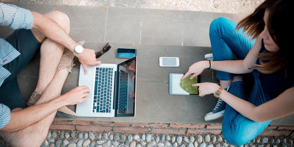 Zwei Personen sitzen sich draußen gegenüber und nutzen Laptop, Tablet und Smartphone für mobiles Internet.