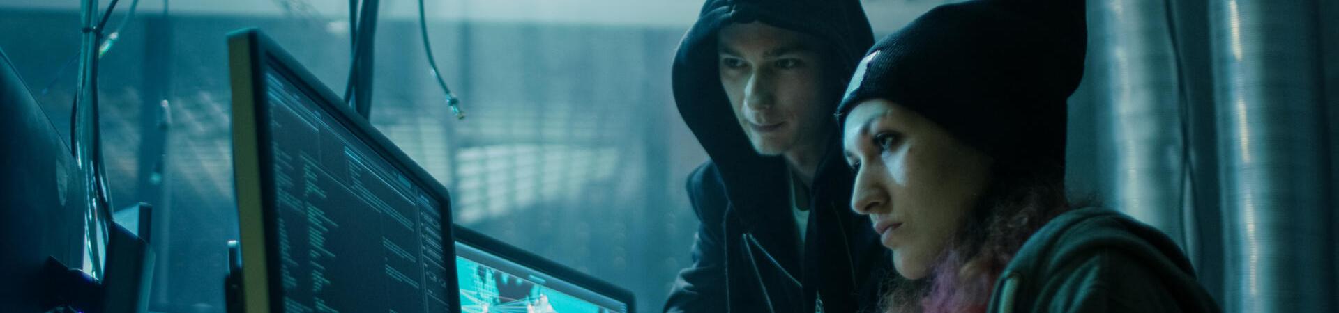 Zwei Personen in Kapuzenpullovern arbeiten an mehreren Computermonitoren in einem dunklen Raum