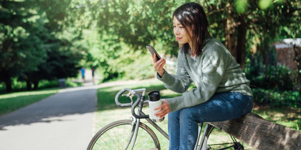 Frau sitzt mit Smartphone und Trinkbecher auf einer Bank neben einem Fahrrad im Park