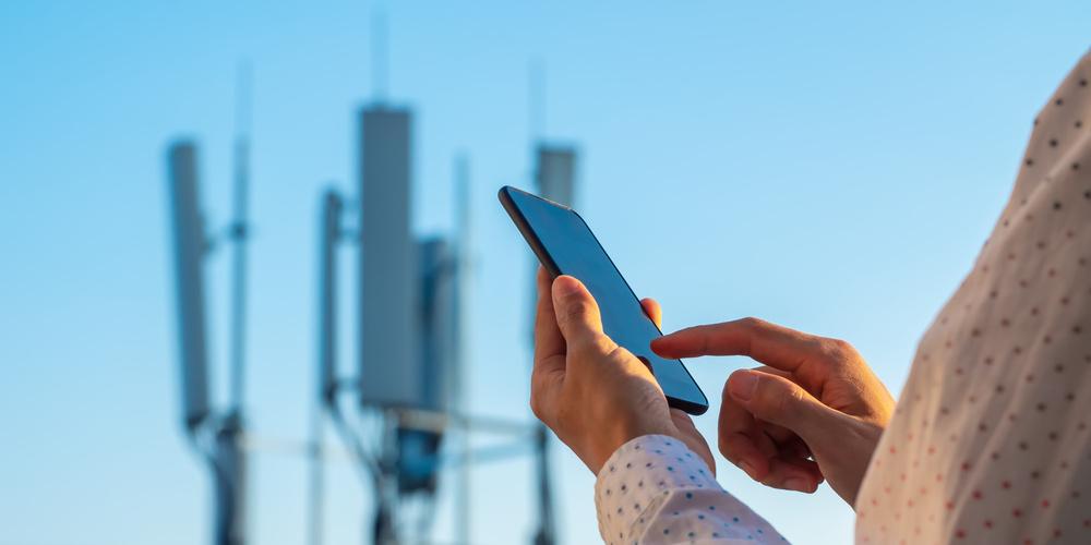 Person nutzt ein Smartphone im Vordergrund, während im Hintergrund unscharf Mobilfunkmasten zu sehen sind.