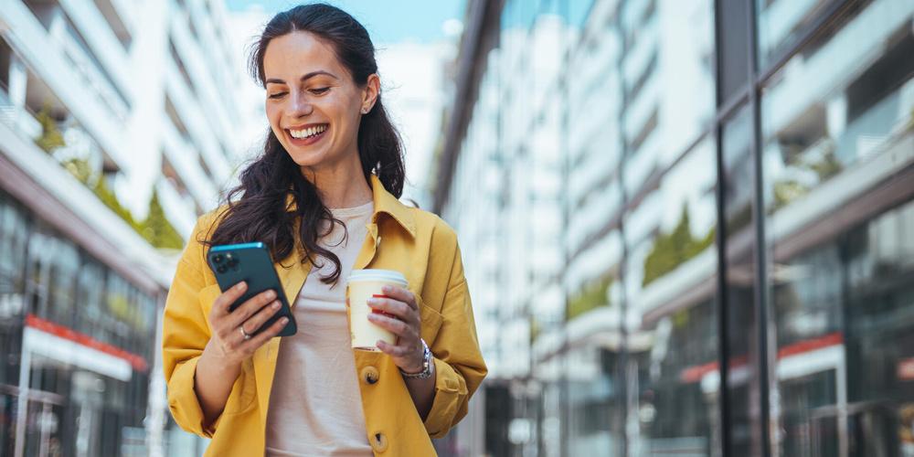 Lächelnde Person hält ein Smartphone in einer Hand und einen Coffee-to-go-Becher in der anderen Hand