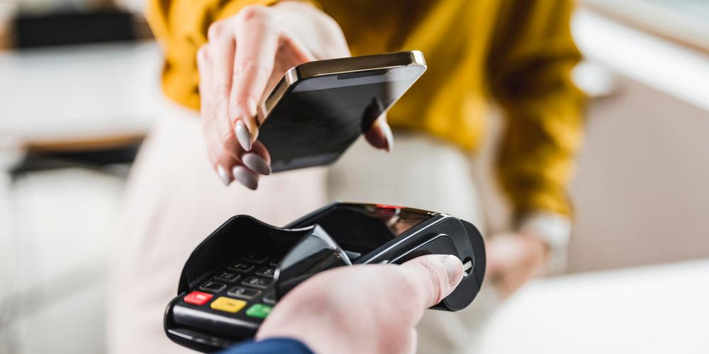 Eine Person hält ein Smartphone über ein Kartenterminal und bezahlt kontaktlos an der Kasse