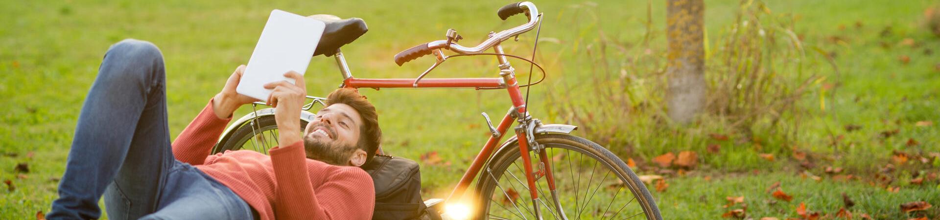 Junger Mann liegt im Wald neben seinem Fahrrad, lehnt an seinem Rucksack und hält ein Tablet in der Hand