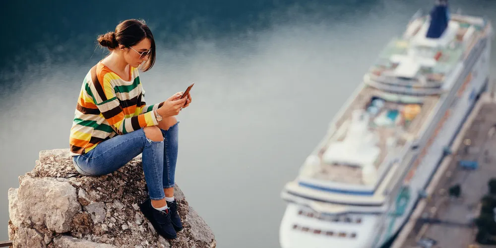 Frau sitzt auf einem Felsen und nutzt ihr Smartphone, im Hintergrund liegt ein Kreuzfahrtschiff im Hafen