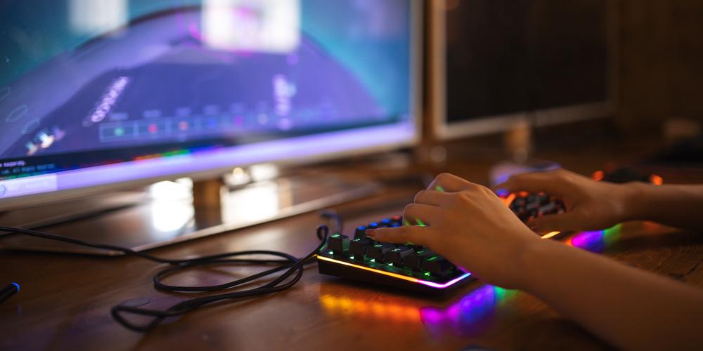 Person spielt an einem Computer mit beleuchteter Gaming-Tastatur vor großem Monitor in dunkler Umgebung