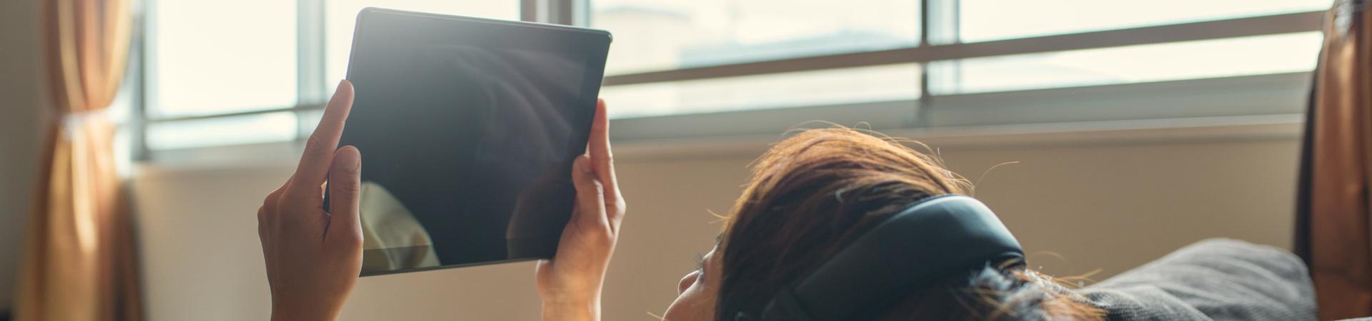 Person liegt mit Kopfhörern und Tablet auf dem Sofa.