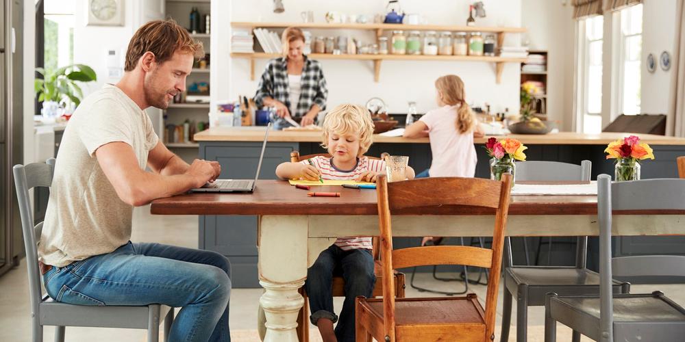 Familie verbringt Zeit in der Küche, ein Erwachsener arbeitet am Laptop, die Kinder malen am Tisch