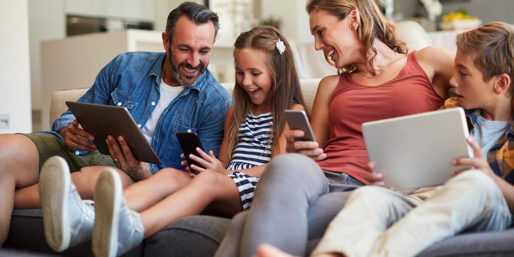 Familie sitzt gemeinsam auf dem Sofa und nutzt verschiedene digitale Geräte