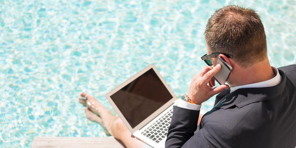 Mann sitzt mit Laptop auf dem Schoß und Smartphone in der Hand am Pool