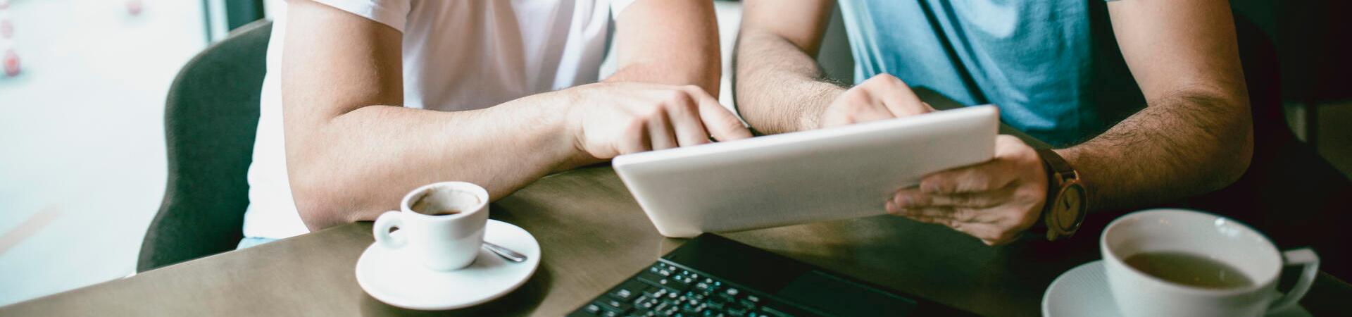 Zwei Männer nutzen gemeinsam ein Tablet mit Tastatur in einem Café