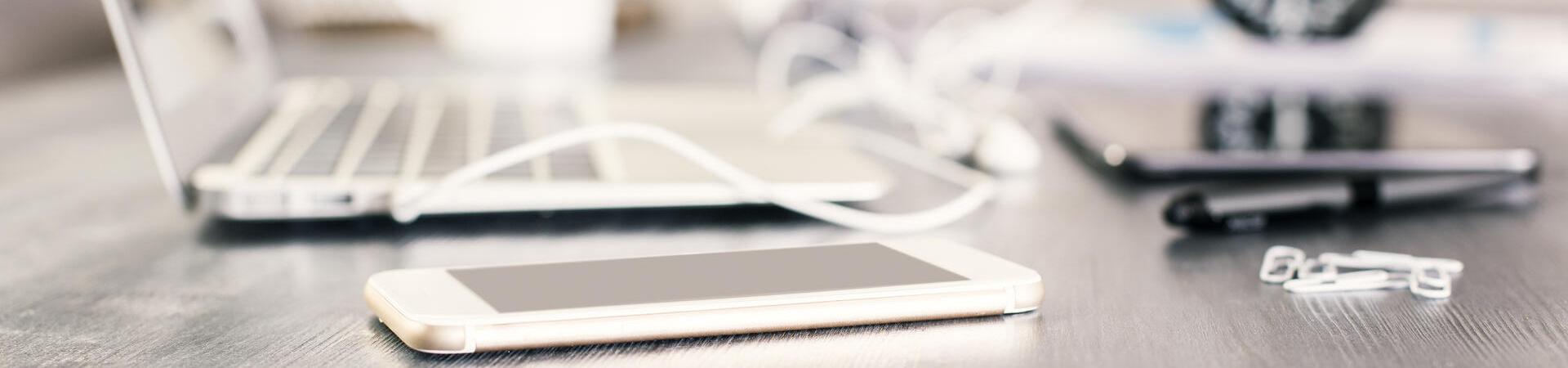 Weißes iPhone auf grauem Tisch mit Laptop und Tablet im Hintergrund