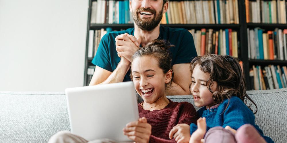 Vater und zwei Kinder sitzen auf dem Sofa und schauen gemeinsam auf ein Tablet