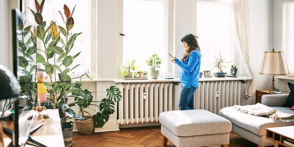Frau steht in einem hellen Wohnzimmer am Fenster und nutzt ihr Smartphone.