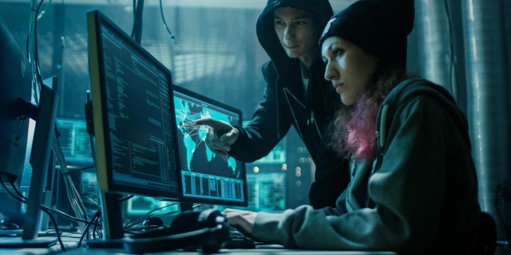 Zwei Personen in Kapuzenpullovern arbeiten an mehreren Computermonitoren in einem dunklen Raum