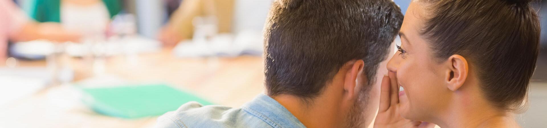 Zwei Personen sitzen in einem mit weiteren Menschen gefüllten Konferenzraum; die eine Person flüstert der anderen etwas ins Ohr.