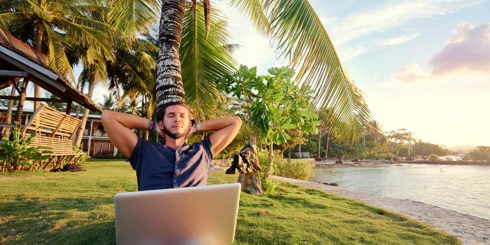 Mann lehnt mit geschlossenen Augen und einem Laptop auf dem Schoß an einer Palme neben dem Wasser