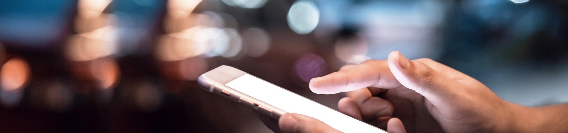 Hände bedienen ein leuchtendes Smartphone vor unscharfem nächtlichem Stadtverkehr mit Bokeh-Lichtern im Hintergrund