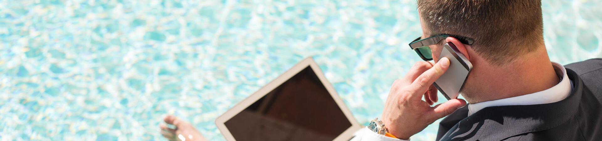 Mann sitzt mit Laptop auf dem Schoß und Smartphone in der Hand am Pool