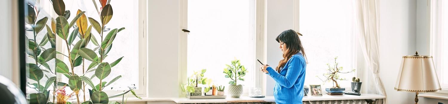 Frau steht in einem hellen Wohnzimmer am Fenster und nutzt ihr Smartphone.