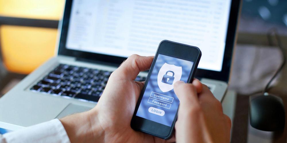 Eine Hand hält ein Handy mit sichtbarer Kennwort-Abfrage; im Hintergrund steht ein Laptop