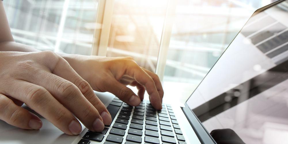 Nahaufnahme von Händen, die auf einer Laptop-Tastatur tippen, neben einem Fenster, mit Sonnenlicht, das auf den Bildschirm reflektiert.