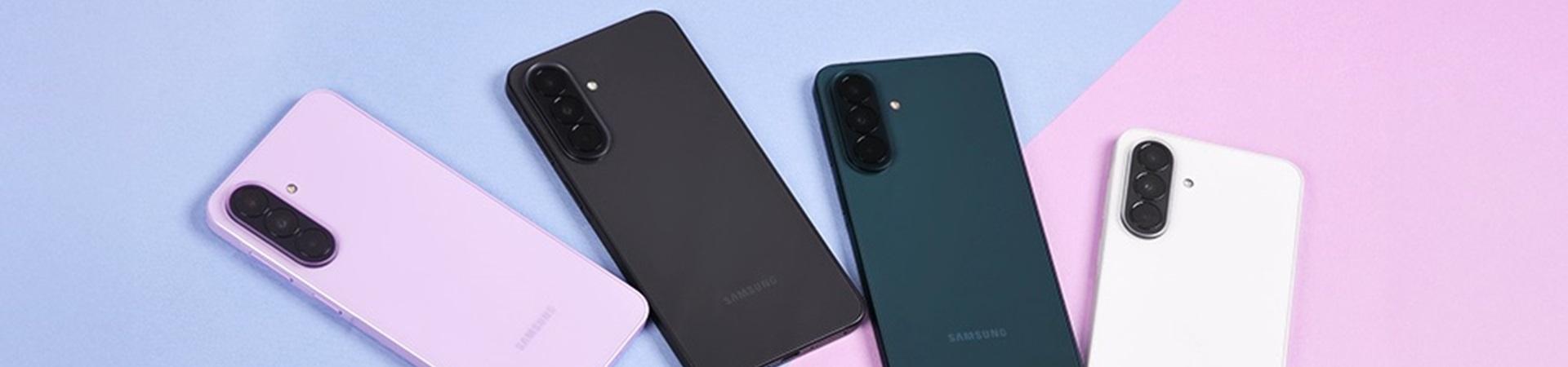 Vier Samsung Galaxy-Smartphones in Rosa, Schwarz, Grün und Weiß liegen nebeneinander auf blau-rosa Hintergrund