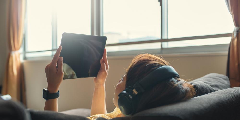 Person liegt mit Kopfhörern und Tablet auf dem Sofa.