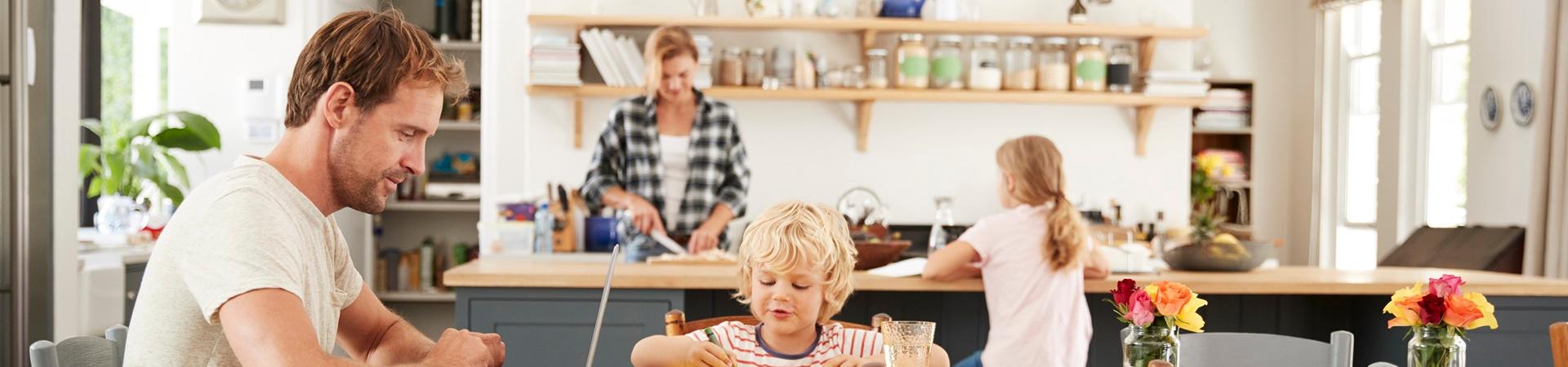 Familie verbringt Zeit in der Küche, ein Erwachsener arbeitet am Laptop, die Kinder malen am Tisch