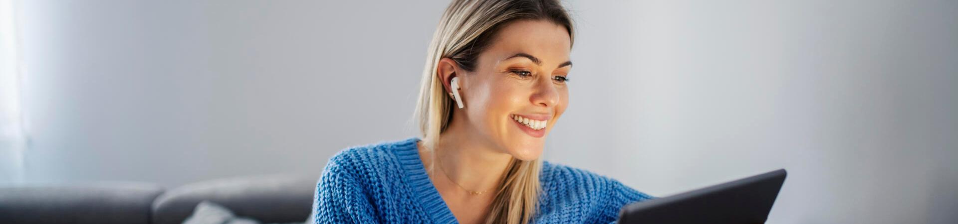Frau hat einen Bluetooth-Kopfhörer im Ohr und schaut lächelnd auf ein Tablet