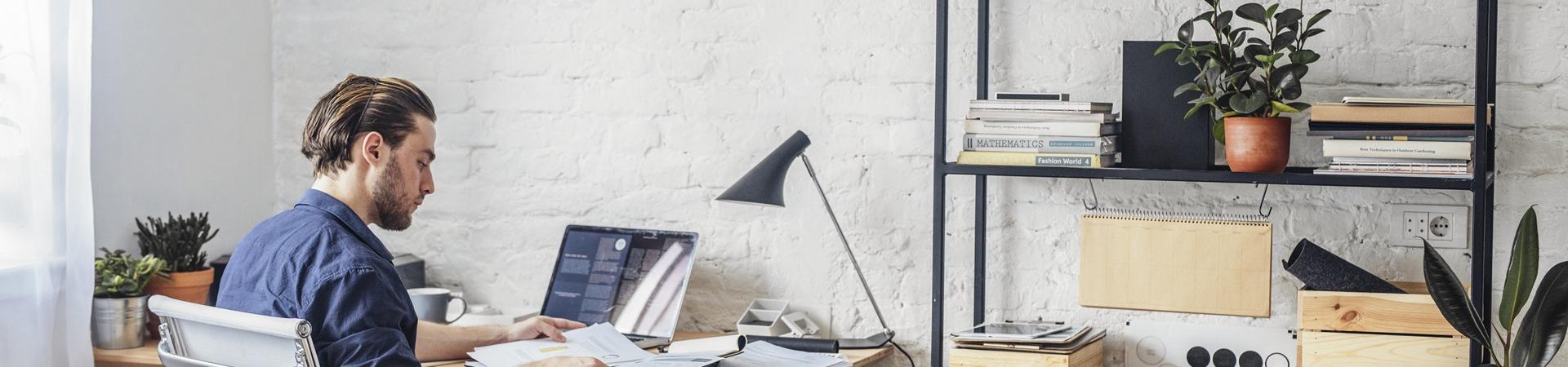 Mann arbeitet an einem Laptop in einem hellen Homeoffice mit Regalen voller Bücher, Ordner und Pflanzen