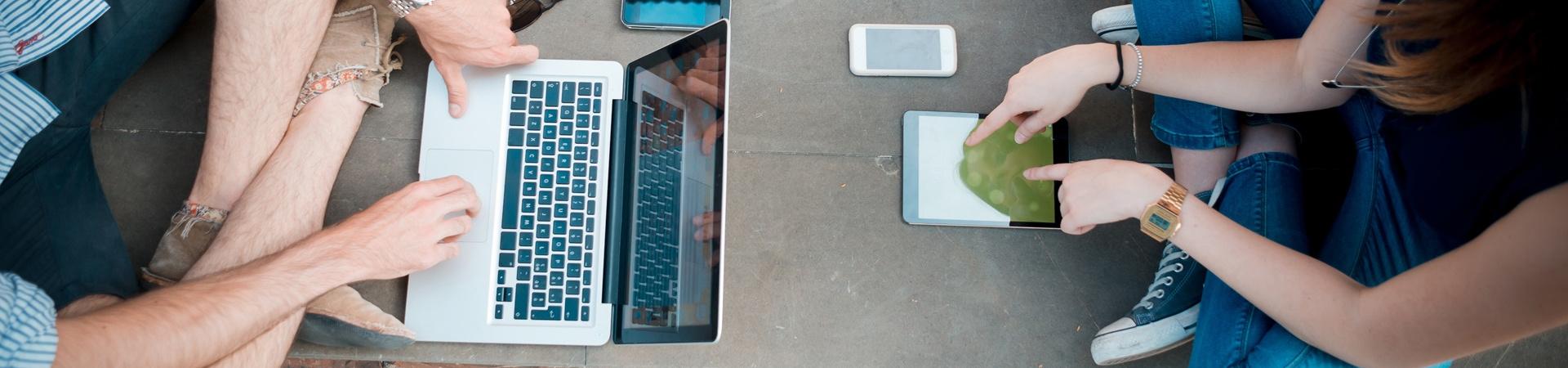 Zwei Personen sitzen sich draußen gegenüber und nutzen Laptop, Tablet und Smartphone für mobiles Internet