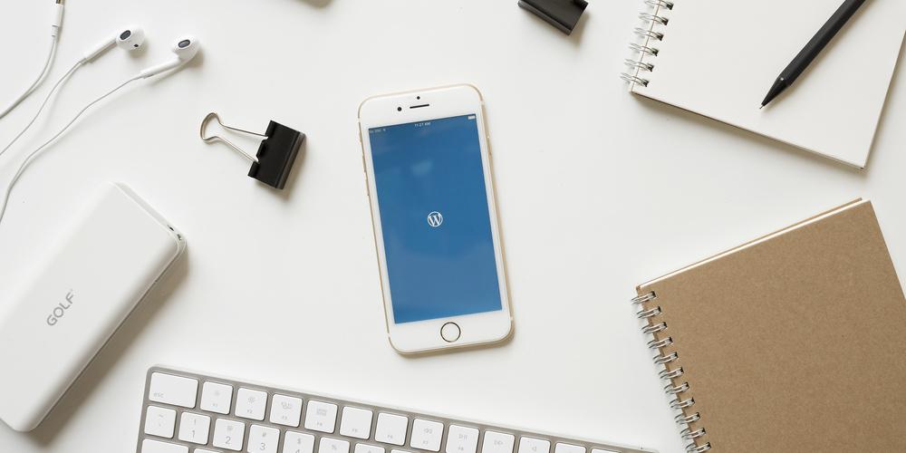 Smartphone auf weißem Schreibtisch neben Tastatur, Notizblöcken, Kopfhörern, Powerbank und Büroklammern