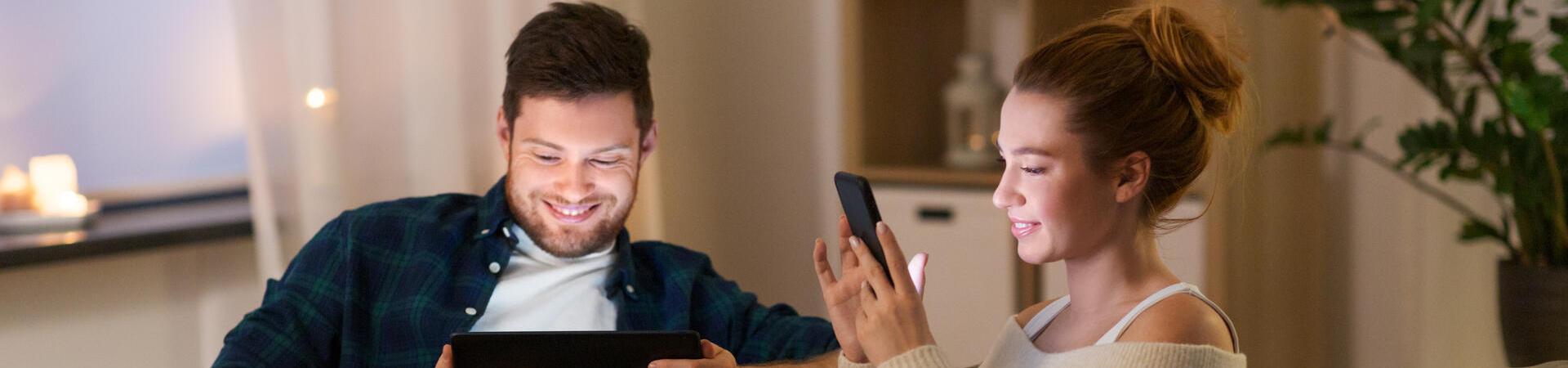 Zwei Personen nutzen Tablet und Smartphone gemeinsam auf dem Sofa