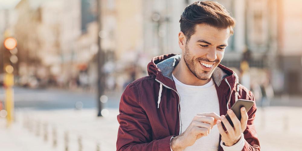 Ein junger Mann steht in einer Einkaufsstraße und blickt fröhlich auf sein Smartphone