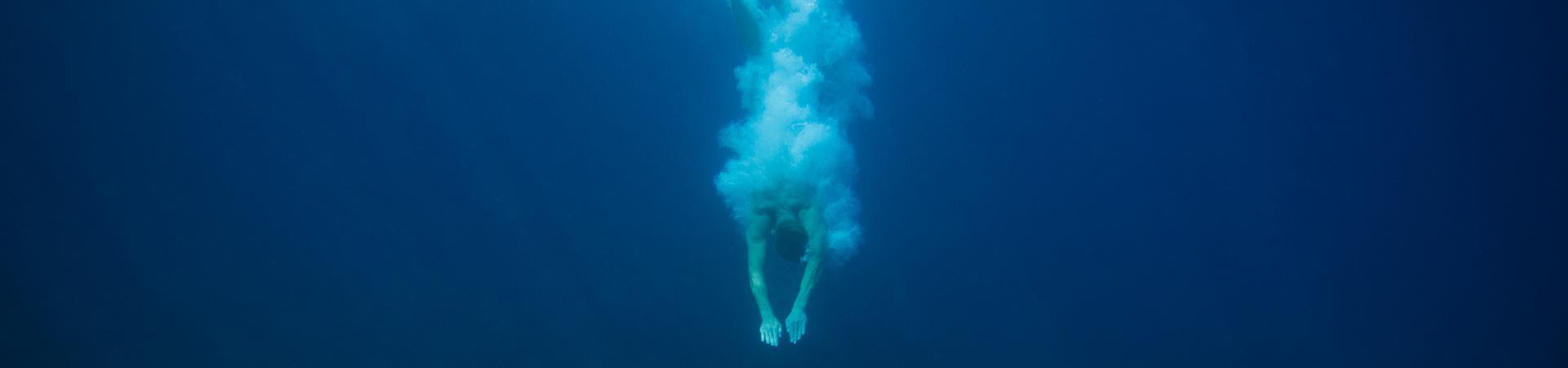Person taucht kopfüber ins offene Meer, unter der Wasseroberfläche mit aufsteigenden Luftblasen vor tiefblauem Hintergrund.