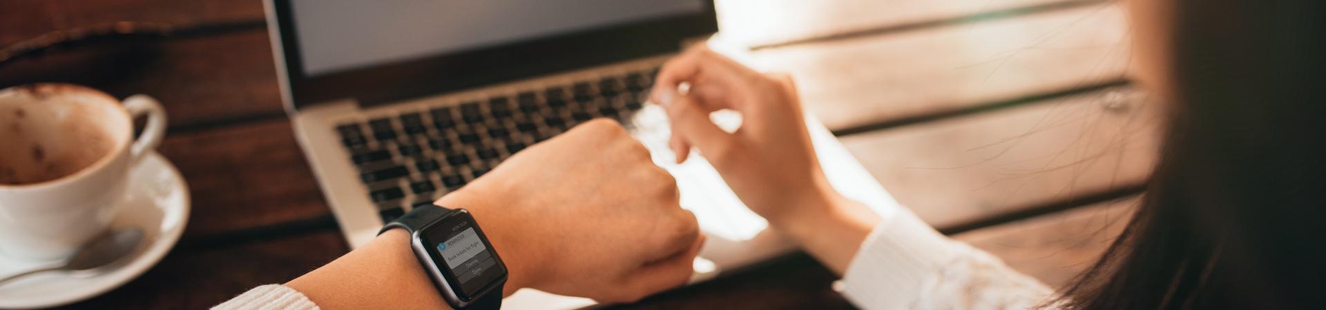 Person arbeitet am Laptop und prüft eine Nachricht auf der Smartwatch.