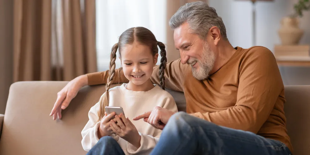 Vater sitzt mit seiner Tochter auf dem Sofa und schaut gemeinsam mit ihr auf ein Smartphone