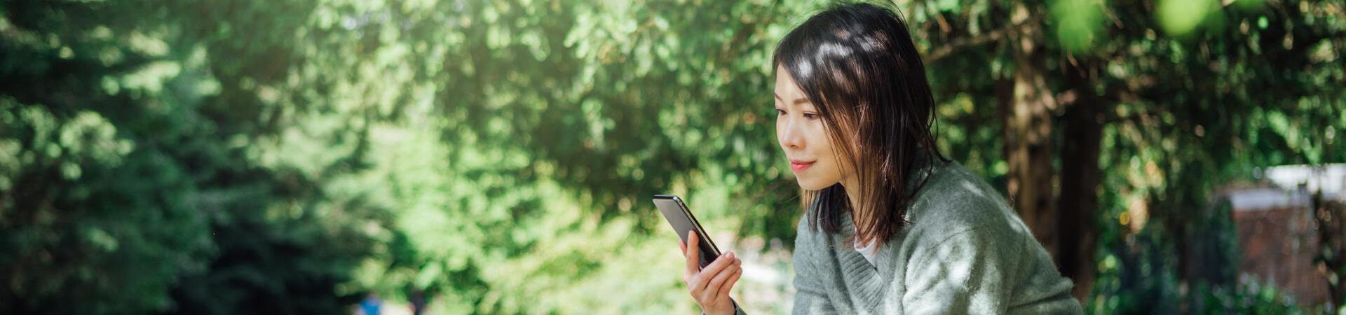 Frau sitzt mit Smartphone und Trinkbecher auf einer Bank neben einem Fahrrad im Park