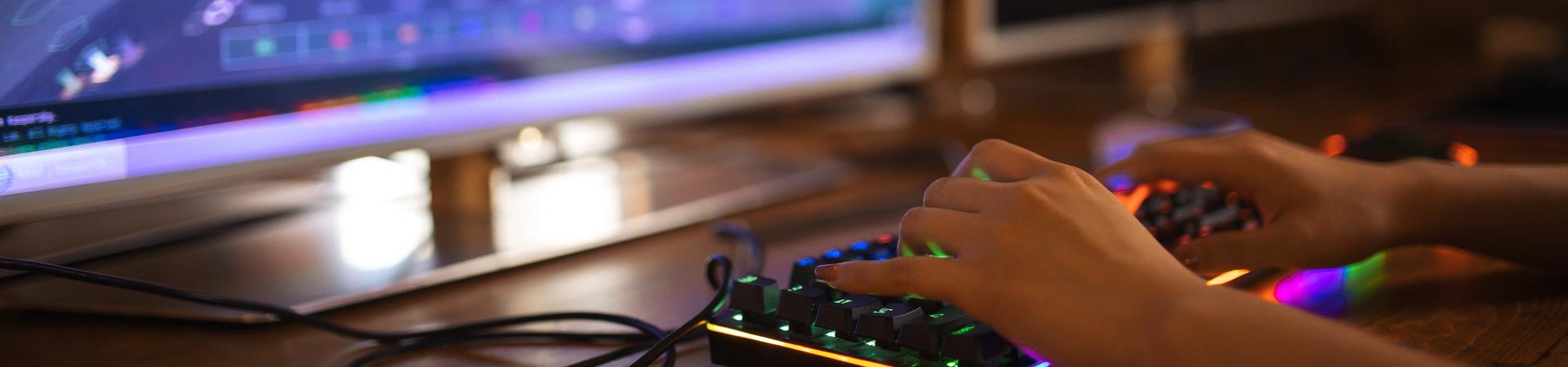 Person spielt an einem Computer mit beleuchteter Gaming-Tastatur vor großem Monitor in dunkler Umgebung