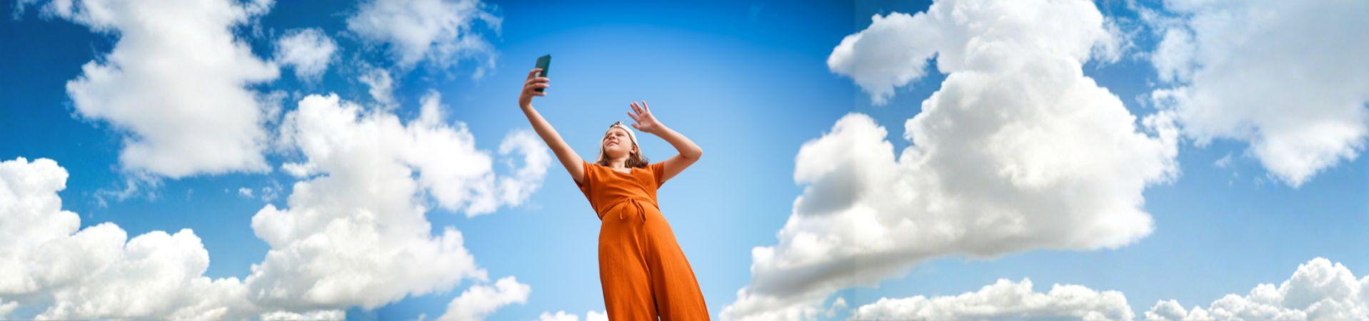 Mädchen macht ein Selfie mit Smartphone vor blauem Himmel mit Wolken.