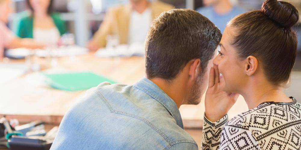 Zwei Personen sitzen in einem mit weiteren Menschen gefüllten Konferenzraum; die eine Person flüstert der anderen etwas ins Ohr.