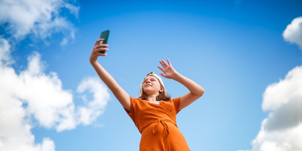 Mädchen macht ein Selfie mit Smartphone vor blauem Himmel mit Wolken.